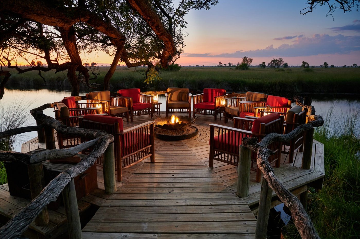 Red armchairs ring the fire pit on a waterside deck at Camp Xakanaxa after sunset