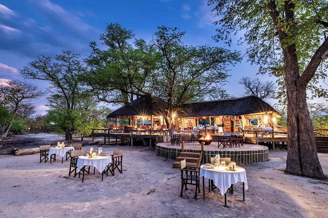 Tables set on the sand before Elephant Pan's lit main area at dusk, north-east of Moremi