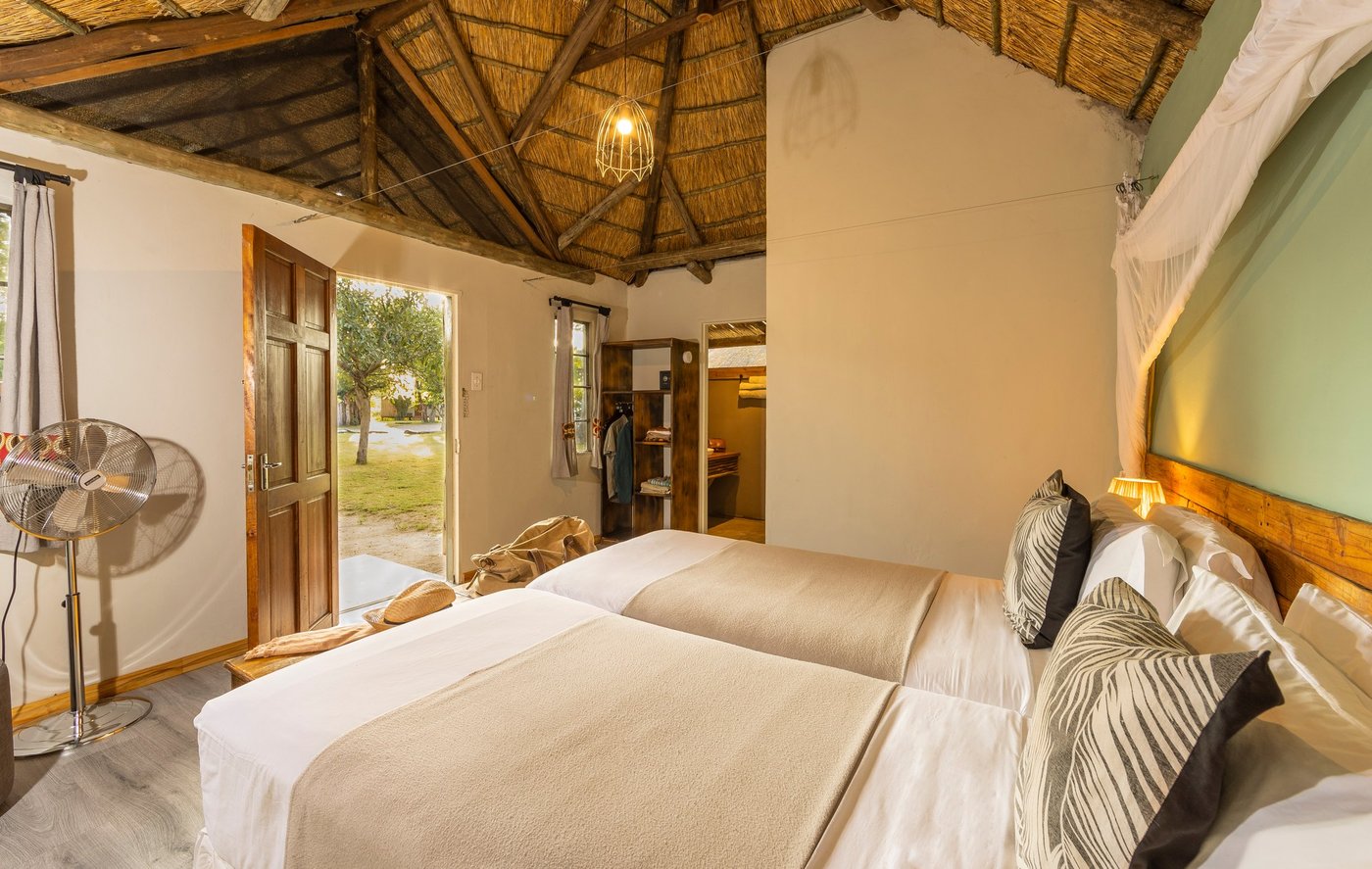 Twin beds under thatch in a Khwai Guest House room, its door open to the lawn