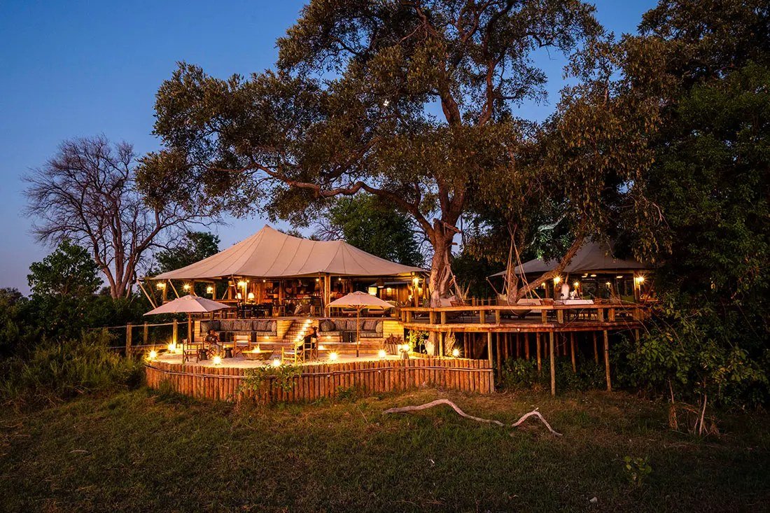 Tuludi's canvas-roofed main area glows under a large tree at dusk, north-east of Moremi